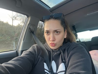 A single female individual is depicted inside a vehicle in Augst, Switzerland. She is seated in what appears to be a front seat, secured by a black seatbelt extending diagonally across her chest and left shoulder. The individual wears a black hooded sweatshirt with a white Adidas trefoil logo on the front. Dark-framed sunglasses are positioned on her forehead, above her dark hair, which is pulled back. Her facial expression features pursed lips, and her eyes are open, directed towards the camera. The vehicle's interior includes a light-colored ceiling with a visible sunroof or panoramic roof structure. A rear seat is partially visible, containing a pink, fluffy object. Through the side window, an outdoor environment is discernible, characterized by trees and green shrubbery along what appears to be a roadside. Bright daylight is evident, with some glare. Distant topographical features, possibly hills or mountains, are faintly visible in the background.