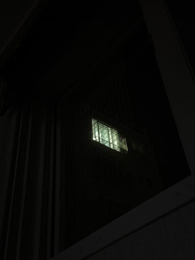 The image shows a nighttime view through a window. The window has a metal grate on the exterior. Inside, a dimly lit building with multiple windows is visible. One section of the building has brightly lit windows, appearing to be offices or rooms. The area outside the window is dark, with some vertical structures that appear to be part of a building or fence. The image was taken in Dongducheon-si, South Korea.