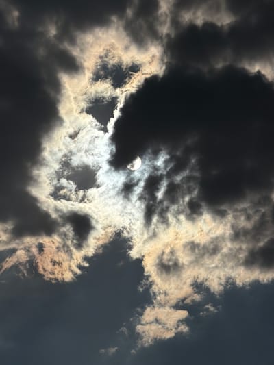 The media file shows a dramatic sky filled with dark, heavy clouds partially obscuring a bright, glowing orb, possibly the sun or moon, near the upper left of the frame. The clouds are a mix of deep grays and blacks, with some edges illuminated by the bright light source, creating a stark contrast. Wispy, lighter clouds are also present, catching the light and adding texture to the scene. The overall impression is one of a turbulent or stormy sky, with the light struggling to break through. There are no people or man-made objects visible, and no text is present. The scene is purely atmospheric and suggestive of a specific weather condition. The location context is Chirchik, Uzbekistan, which could imply the type of cloud formations or lighting conditions common to that region. The time of day is ambiguous, but the strong contrast between light and shadow might suggest either dawn, dusk, or a day with heavy cloud cover. The mood is somewhat ominous due to the dark clouds, yet also awe-inspiring due to the powerful light source piercing through.