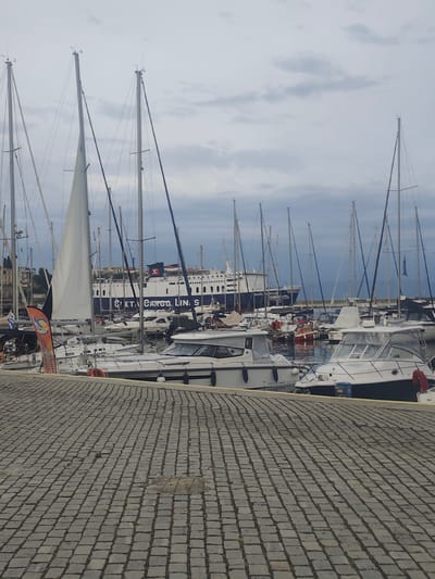 The image depicts a harbor in Kavala, Greece, with numerous boats docked and a large ferry in the background. The foreground is dominated by a cobblestone pathway leading towards the water. Several sailboats, characterized by their tall masts and rigging, are prominently featured, alongside smaller motorboats. One sailboat in the mid-ground has a partially unfurled white sail. In the background, a large ferry, identified by the text "AGIOS GEORGIOS" and "AGIOS GEORGIOS" on its side, is docked. The ferry has a blue and white hull and a red "K" logo. The sky is overcast, suggesting a cloudy day. No people are clearly visible in the image. The overall scene is one of a busy maritime port.