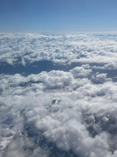 The image presents a stunning aerial view, likely captured from an aircraft, showcasing a vast expanse of clouds and mountainous terrain. The left side of the frame is dominated by a clear, vibrant blue sky, gradually becoming hazier towards the approximate "horizon" line that runs vertically down the image.
To the right of this demarcation, a sea of dense, fluffy white cumulus clouds stretches across the landscape. These clouds are organized into striking, elongated, and largely parallel rows, a meteorological phenomenon often referred to as cloud streets, which recede into the distance, conveying depth. Bright sunlight illuminates the tops of these clouds, creating soft highlights and casting subtle shadows within and beneath them, indicating it is daytime, likely late morning or early afternoon.
Peeking through and above the extensive cloud cover are the rugged, snow-capped peaks of mountains. These dark, formidable landforms are scattered across the right half of the image, with significant amounts of snow visible on their higher elevations. Patches of darker ground or valleys are occasionally visible between the cloud formations and mountain ranges, though specific details are obscured by the distance and cloud density.
The scene evokes a sense of vastness and tranquility, capturing the natural beauty of a high-altitude landscape. There are no people or discernible text visible in the image. Given the location context of Electoral Area A, Canada, the view is over a mountainous region, consistent with the majestic peaks and extensive cloud formations often found in such areas.
