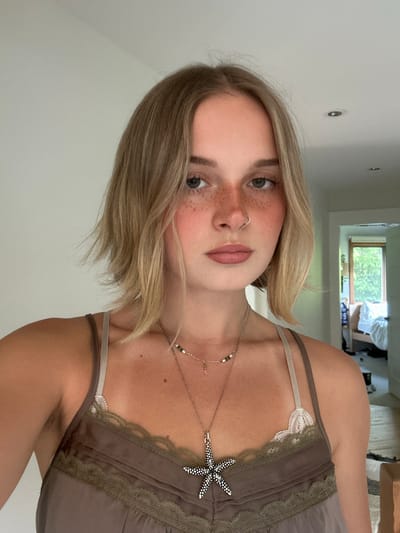 A young adult female is captured in a close-up, head-and-shoulders, likely selfie-style portrait, within an indoor residential environment. She has short, light blonde hair, styled in a bob, and fair skin with numerous freckles across her nose and cheeks, enhanced by a visible red blush. A silver hoop nose ring is present in her left nostril. She wears a brown or taupe camisole with dark lace trim and thin straps; a white lace underlayer is also visible on her right shoulder. She accessorizes with two necklaces: a shorter one with small beads and a delicate charm, and a longer one featuring a prominent silver and black speckled starfish pendant. Visible armpit hair is present under her left arm. The background is a plain, light-colored wall, with a partial view of an open doorway on the right leading to another room. Through this doorway, a window reveals abundant green foliage outdoors. The specific location in the city of Squamish, Canada, cannot be visually confirmed from the details present in the image.