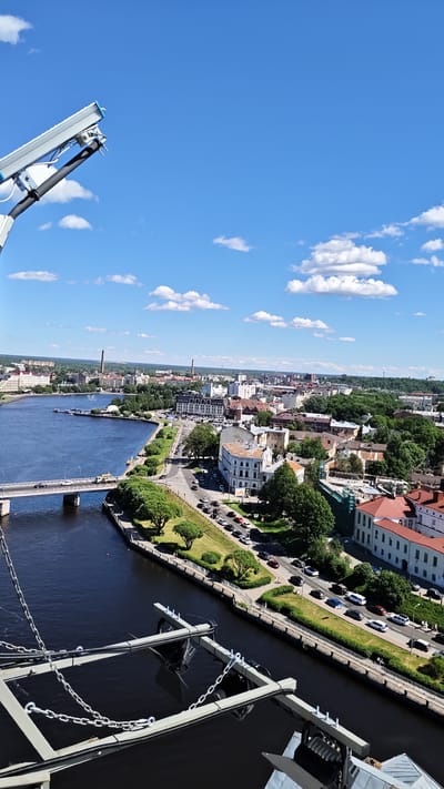 An aerial view of the city of Vyborg, Russia, is captured on a clear day with scattered white clouds against a bright blue sky. The scene focuses on a wide, dark blue river that cuts through the urban landscape. A prominent bridge with a light gray railing spans across the river, with cars visible on its surface.
Along the riverbank, a tree-lined promenade and a road are visible, with a steady flow of traffic, including cars and what appears to be a bus. Various buildings, characterized by red-tiled roofs and light-colored facades, line the riverside and extend into the city. Some structures are older and more ornate, while others are more modern. Green trees and landscaping are interspersed throughout the urban environment.
In the foreground, a portion of an industrial or structural element is visible, featuring a gray metallic framework with hanging chains and cables. This element appears to be part of a large machine or piece of equipment, possibly a crane or a lighting rig, extending into the frame from the bottom. In the distance, more of the city stretches towards the horizon, with several industrial chimneys piercing the skyline. The overall impression is a bustling city viewed from a high vantage point on a sunny day.