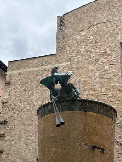 The image displays a close-up, low-angle view of a fountain located in Foligno, Italy, set against an ancient building facade. The central element is a cylindrical, light brown concrete fountain column, marked by water stains and textured surfaces. This column supports a darker, circular basin from which water is actively cascading over its rim. Two patinated bronze statues of childlike figures are positioned on the basin's edge. The figure on the left holds a long, double-barreled horn pointing downwards, while the figure on the right holds a small, round object with attached lines. Both figures are depicted wearing distinctive, wide-brimmed hats or headdresses. A dark-feathered pigeon is perched atop the headdress of the rightmost bronze statue. The background is dominated by a multi-story building constructed of light-colored, irregularly shaped bricks, exhibiting variations in tone and texture consistent with age. An overcast grey sky occupies the upper left portion of the frame, bisected by a thin, dark wire.