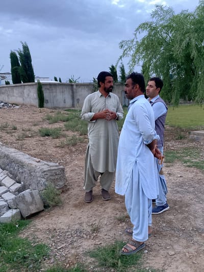 Three men are engaged in a conversation outdoors under an overcast, grey sky, suggesting late afternoon or an entirely cloudy day. The setting appears to be a semi-urban or rural area in Qila Saifullah, Pakistan.

The man on the left, facing right, wears a light olive-green shalwar kameez and dark slip-on shoes. He has a beard and mustache and gestures with his hands, which are clasped together. The man in the center, partially visible, is seen from the side, looking towards the man on the left. He is dressed in a light blue shalwar kameez and dark sandals. He also has a beard and mustache, and his hands are clasped behind his back, holding a string of light brown prayer beads (tasbih). The third man, on the far right and mostly seen from behind, wears a light blue shirt with a dark grey or blue sleeveless vest over it, and dark sneakers. He is also looking towards the other two.

The ground beneath them is unpaved, composed of dry, light brown soil with scattered small stones, patches of sparse green grass, and some dry vegetation. To the immediate left, a low concrete structure, possibly a planter or part of a foundation, is visible, with concrete blocks stacked beside it. A tall concrete wall stretches across the mid-ground. Behind this wall, several tall, slender evergreen trees (likely cypress) stand, and glimpses of white buildings are visible. On the right side, a large weeping willow tree with vibrant green foliage provides some shade, and a section of neatly maintained green lawn can be seen further back. The scene depicts a casual, yet seemingly earnest, discussion among the men in this outdoor environment. No visible text is present.