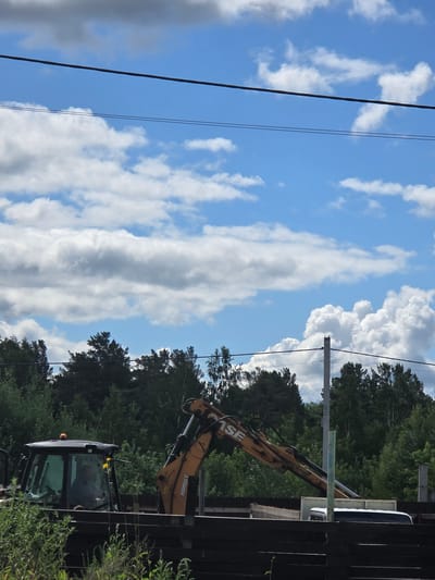A backhoe loader is parked in an outdoor area with trees and a cloudy blue sky. The backhoe, which is yellow and black, has its arm extended. A person is visible inside the cabin of the backhoe. The area appears to be under construction or renovation, with a wooden fence and some construction materials in the foreground. The location is identified as Ermolaevo, Russia.