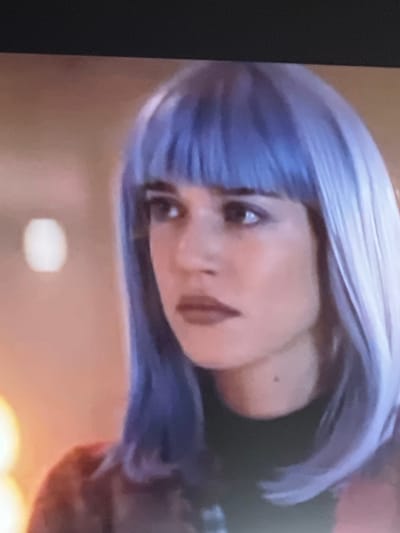 This media file is a close-up, vertically oriented image of a person's face and hair. The subject appears to be female, with light purple or blue hair styled in a bob with bangs. Her face is turned towards the viewer, with her eyes looking directly ahead. She has dark eyebrows, and her lips are painted a reddish-brown color. There is a small, dark mole visible on her cheek. The lighting is somewhat dim, with a soft glow emanating from an unseen source, suggesting an indoor setting. The background is blurred, with warm tones of orange and yellow. The overall mood of the image is somewhat ambiguous due to the angle and lighting, but the direct gaze of the subject could be interpreted as confident or introspective. There is no discernible text in the image. The location context provided is Baošići, Montenegro, but there are no visual cues within the image to confirm this specific location.