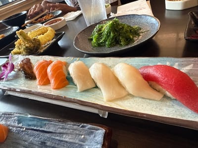 The image shows a table at a restaurant in Orlando, Florida, United States. A variety of Japanese dishes are arranged on the table, including a sushi platter with eel, salmon, white fish, and tuna nigiri. There is also a plate of seaweed salad and a dish of tempura. People are visible in the background.
