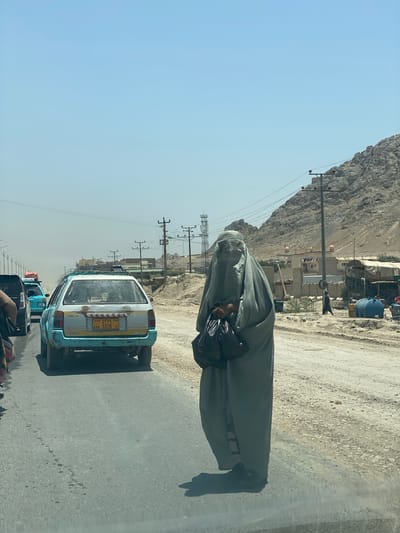 In an outdoor, daytime scene in the city of Kokran, Afghanistan, a woman wearing a burqa stands on the side of a road. She is holding two black plastic bags. The road is dusty and appears to be a busy thoroughfare, with several cars visible in the background, some of which are also on the road. The surrounding environment includes barren hills, scattered buildings, and power lines. The overall impression is one of a rural or semi-urban area in a dry, mountainous region.