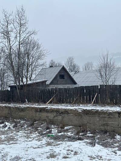 This media file captures a stark, wintry rural scene in Yasinya, Ukraine. The central subject is a rustic, weathered wooden building, possibly a barn or an old house, with a steep, corrugated metal roof and a small, dark, square window on its gable end. The building is situated behind a tall, dark wooden fence, which has several diagonal wooden supports leaning against it.

In front of the fence, a muddy trench or ditch spans across the mid-ground. Beyond the trench, the ground is partially covered in snow, with patches of dry, sparse vegetation visible. A small, green plastic bottle lies on the snow-covered earth towards the mid-right. The sky is uniformly light gray and overcast, suggesting a cloudy, cold winter day. Several bare deciduous trees with intricate branch structures are prominent, both in the foreground and receding into the background, emphasizing the winter season. No people or specific activities are depicted in the static scene, and no visible text is present. The overall impression is one of quietness and rustic charm, characteristic of a winter landscape in a rural area.