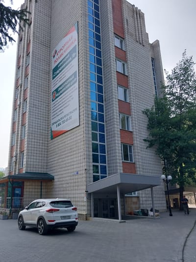 The image shows a multi-story building with a sign that reads "КУРОРТНАЯ ПОЛИКЛИНИКА" (Resort Polyclinic). The building is constructed with light-colored bricks and features several windows, some with blue reflective glass. A white SUV is parked in front of the building. The sign also lists services offered, including "ОБЩЕКУРОРТНЫЙ МЕДИЦИНСКИЙ ЦЕНТР" (General Resort Medical Center). A phone number is visible on the sign: 8 804 700-65-50. A street sign in the background appears to read "САНАТОРИЙ" and a number, likely indicating a street name. The environment suggests an outdoor scene in a developed area, possibly a resort town. The overall impression is a functional, public building with a focus on medical services.