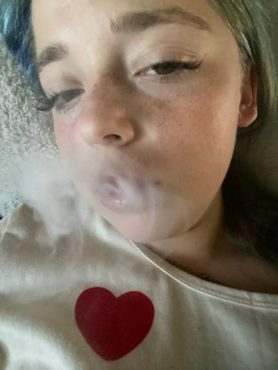 The image is a close-up, head-and-shoulders shot of a young person with light skin, freckles, and light-colored hair, some of which has blue streaks. The person is wearing a white t-shirt with a red heart graphic. They appear to be exhaling a cloud of white vapor, obscuring their mouth and nose. The background is a textured, light-colored surface, possibly a blanket or bedding. The lighting is soft and appears to be coming from above. The image was taken indoors. The location in Nova Praha, Ukraine, is not discernible from the image.