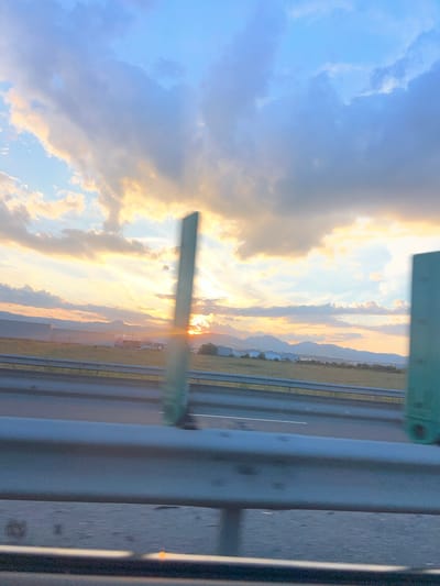 The image depicts a sunset scene captured from a moving vehicle on a highway, likely near Oiejdea, Romania, given the context. The sky is a prominent feature, filled with a mix of light blue, white, and golden-orange clouds. Rays of sunlight pierce through the clouds, creating a dramatic effect. In the distance, a range of mountains is visible, silhouetted against the vibrant sky. Below the mountains, a flat landscape stretches out, dotted with industrial buildings, some appearing as large, elongated structures and others as smaller complexes. A body of water, possibly a lake or river, reflects the light of the setting sun, though this is difficult to discern clearly due to the image quality. The foreground is dominated by a highway guardrail, which is metallic and light gray, partially obscuring the view of the road ahead. The road itself appears wet, with white lane markers visible. Two tall, rectangular objects, possibly signs or roadside markers, are positioned along the highway; one is in the middle ground and another is on the right side of the frame, both appearing to be a muted green color. The overall impression is one of a serene, yet somewhat industrial, roadside landscape at dusk. There are no people or animals visible in the image.