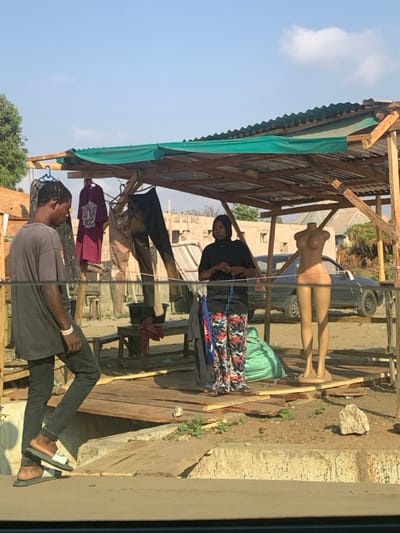 An outdoor scene in Abobo, Nigeria, depicts an informal roadside clothing stall under a clear blue sky with minimal cloud cover.
The stall features a makeshift structure comprising wooden posts supporting a corrugated metal roof, partially covered by a green tarp. A raised wooden platform, constructed from planks, serves as the floor of the stall.
In the left foreground, slightly below the stall's platform, an adult male with dark skin and short dark hair stands, facing right and looking downwards. He wears a dark, short-sleeved t-shirt, dark pants, and stacked sandals (dark sandals with white-strapped lighter sandals on top). A light-colored wristband is visible on his left wrist.
Inside the stall, on the wooden platform, an adult female with dark skin stands centrally, facing slightly right. She wears a black long-sleeved top, a dark headscarf, and wide-legged pants with a red, white, and blue pattern. Both her hands hold a blue chain-like object at chest level. To her right, a light brown, headless female mannequin, comprising a torso and legs, stands upright on the platform. A crumpled green bag rests on the platform behind the mannequin. Several pieces of clothing, including t-shirts and trousers, hang from the stall's wooden framework.
In the background, a dark-colored sedan is parked to the right of the stall. Further back, unfinished masonry buildings and sparse vegetation are visible on dusty, unpaved ground. A wooden bench or table with a dark container on it is present in the middle ground behind the man. The immediate foreground consists of a concrete curb or edge.