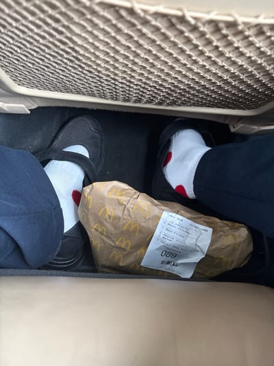 The image depicts the footwell of a vehicle located in Staffordshire Moorlands, United Kingdom. Two individuals' lower legs and feet are visible on a dark floor surface. The individual on the left wears dark blue trousers, a white sock with a red heart design near the ankle, and a black Mary Jane-style shoe with a strap. The individual on the right wears dark trousers, a white sock with a red heart design, and a black slip-on shoe. Between their feet rests a brown McDonald's paper bag, adorned with multiple yellow 'M' logos. A white label affixed to the bag details a "Take Out" order (Order: 089) placed on "08/03/2020 12:49". The order contents listed are "3 x Selects", "1 Mayo Chicken", "1 Med Fries", "2 Apple Juice", "2 Water", and "1 Smoky BBQ Dip". A light-colored mesh pocket, typically found on the back of car seats, is visible in the upper portion of the frame. The subjects are stationary within the vehicle, and the McDonald's order is positioned on the floor, indicating recent acquisition or impending consumption.