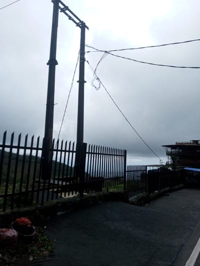 This image captures an outdoor scene in Bangtsho, Bhutan, under an overcast, gray sky. In the foreground, a dark asphalt road occupies the lower right quadrant, extending upwards and slightly to the right. Adjacent to the road on the left, on a small patch of dark ground, two reddish-brown, spherical objects are visible next to a dark, woven fragment. A dark metal picket fence with pointed tops runs across the mid-ground, supported by a low, dark stone or concrete wall. Behind the fence, two tall, dark utility poles with a connecting horizontal crossbar stand prominently. Multiple dark wires are affixed to these poles, stretching across the frame, with one segment appearing loosely coiled to the right of the left pole. Beyond the fence, distant hillsides are covered in dense green vegetation, with structures faintly visible in a valley below. On the far right, a multi-story building featuring dark facades and prominent balconies is visible, situated on elevated terrain, with a blue tarp partially visible beneath it. No individuals are present in the scene.