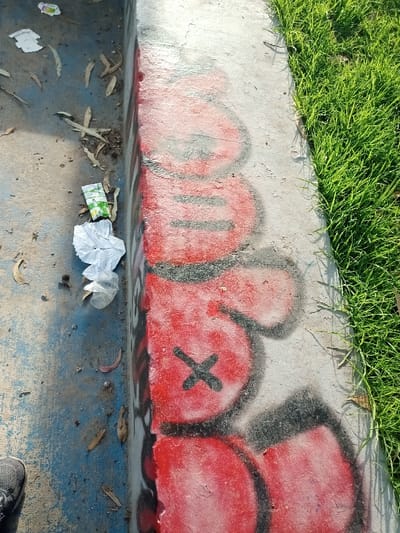 A concrete curb divides blue pavement with litter from green grass. Red and gray graffiti of cartoon faces adorns the curb's side. One face displays an "X" for an eye. Leaves, a juice box, and crumpled paper lie scattered on the pavement. A shoe is visible at the bottom left corner. The scene is captured outdoors in daylight.