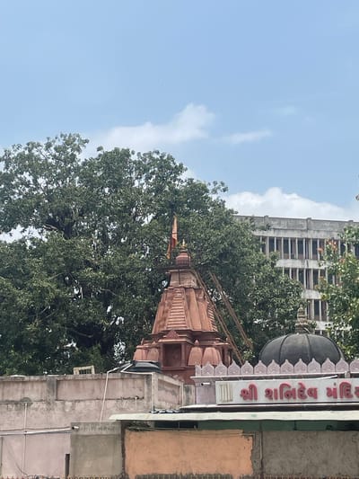 The image shows a religious structure with a prominent dome and a tiered spire, likely a temple, located in Ahmedabad, India. The structure is made of stone and appears to be of traditional Indian architecture. A large tree with dense foliage is situated behind the temple, and a multi-story building with a grid of windows is visible in the background. The sky is partly cloudy, with a few scattered clouds against a pale blue backdrop. The scene appears to be captured during the daytime. There are no people or active interactions visible in the image.