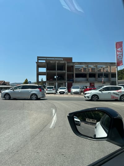 A bright, sunny day unfolds in Mrčevac, Montenegro, with a clear blue sky overhead. The scene captures a street intersection in front of an unfinished concrete building, its skeletal structure suggesting ongoing construction. Several cars are parked or in motion: a silver station wagon is visible on the left, while a white hatchback with red stripes and the word "BAR-KOC" emblazoned on its side is in the foreground. Behind it, a red car and a white sedan are parked. A red banner with the word "IDEA" vertically printed on it hangs from a pole on the right side of the frame. In the foreground, a car's side mirror reflects a white sedan and some wooden structures behind a barrier on a road. The road surface is asphalt, marked with white lines, and shows signs of tire marks. The overall impression is one of a typical urban scene on a warm day.
