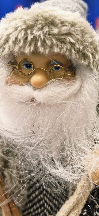 The image provides a vertical, close-up view of a static decorative figurine, located in Vendin-le-Vieil, France. The subject depicts the head and upper body portion of a figure characterized by a light beige face with painted blue eyes accented with white glitter, a small, peach-toned nose, and circular, gold-colored spectacles positioned over the eyes. The figure wears a voluminous, white artificial fur beard, extending from below the nose and covering the lower face and upper chest area. Fine, translucent, hair-like fibers are visible across the beard and spectacles. A grey artificial fur hat, featuring a distinct lighter grey band, is worn on the head. Below the beard, a garment with a black and light grey or white chevron pattern is partially visible. A light beige, coarse-textured braided rope runs vertically along the lower right edge of the frame. The background is an out-of-focus blue field at the upper left, transitioning to darker tones. Small, reddish-brown particulate matter is embedded within the white fur beard, alongside scattered silver glitter fragments. No human figures are present, and no actions or interactions are depicted.
