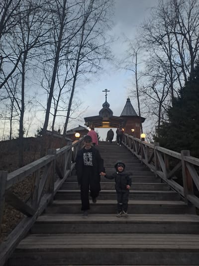 The image depicts a family walking on a wooden bridge leading to a church. The church has a distinct architectural style with a dome and a cross atop it. The sky is overcast, and bare trees flank the bridge. The location is Vzglyadnevo, Russia. The people are on the bridge, walking toward the church.