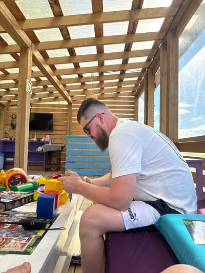 A man with a beard and sunglasses is sitting at a white table outdoors. He is wearing a white t-shirt and white Puma shorts. He is holding building blocks and appears to be playing with them. The table is covered with toys, a board game, and a walkie-talkie. In the background, there is a wooden structure with a clear roof, a television, and shelves with more toys. The scene is lit by natural sunlight. The location is in the city of Zamosh'e (Zaklinskoe S/P), Russia.