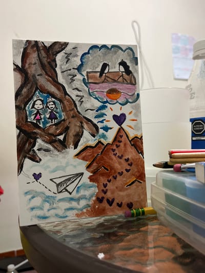 The image shows a small, hand-painted canvas depicting a surreal landscape. On the left, a large, gnarled tree trunk frames two stick-figure girls in dresses, one pink and one purple. Emerging from the tree, the landscape shifts to a mountain range dotted with small hearts leading up to a large purple heart at the summit. Above the mountains, a thought bubble shows two birds on a perch with a sunset. A paper airplane flies through a sky with choppy blue clouds and lightning bolt shapes. The canvas is placed on a glass-topped table, reflecting the scene. Next to the canvas is a clear plastic container filled with art supplies, including pencils and pens. A white bottle with a blue label that reads "ALTO EN AZUCAR" (High in Sugar) is also visible behind the canvas. The background appears to be a white wall with some faded posters. The overall scene suggests an indoor setting, possibly a craft area or workspace. There is no indication of the location being in San Luis, Peru.