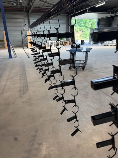 The image shows a close-up, low-angle view of black metal racks and hangers suspended from an overhead track system. The scene appears to be an industrial setting, possibly a warehouse or manufacturing facility, with a concrete floor visible below. The racks are arranged in rows and are designed to hold objects, indicated by the numerous small, hook-like protrusions extending from them. Some of the racks are empty, while others have faint shadows cast on the floor, suggesting they might be in use or have recently been used.
In the background, a large opening reveals lush green trees and foliage, implying the presence of an outdoor area or a large window. The lighting suggests it is daytime, with the light source coming from the open area. The overall impression is one of a functional, organized space, likely involved in production or storage. There are no people visible in the image. The context provided places this scene in Mol, Belgium.