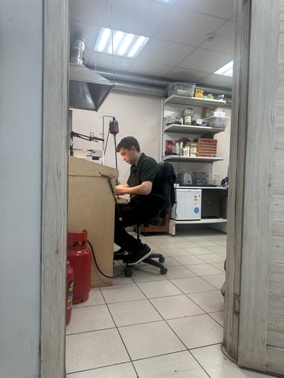 This is a vertically oriented, eye-level photograph taken indoors. The main subject is a young man, dressed in a dark green t-shirt and black pants, kneeling on a black rolling office chair. He is actively engaged in some sort of work or repair, leaning over a wooden workbench. His hands are occupied with tools and equipment mounted on the workbench.
The setting appears to be a workshop or a back room of a store. Behind the man and the workbench, there are shelves stocked with various items, including bottles, containers, and what looks like lab equipment. A small white refrigerator is also visible. To the right of the workbench, there are two red propane tanks. The floor is tiled with light-colored square tiles. Overhead, fluorescent lights illuminate the space. The overall impression is of a functional, somewhat cluttered, work environment.
The image seems to have been taken during daylight hours, judging by the artificial lighting and the lack of external light sources. There are no clear indications of weather or time of day beyond that. The man's expression is focused on his task, suggesting concentration.
Visible text includes what appears to be a label on the smaller propane tank, although it is not legible. A blue sticker with a circular symbol is on the refrigerator.
The location context provided is Tver, Russia.