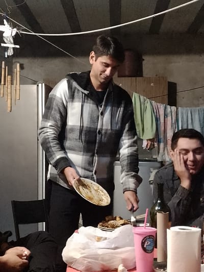 A man wearing a grey and white plaid hooded jacket and black pants is standing at a table cutting food with a fork. He's holding a dirty plate in his left hand. Another man is seated at the table to his right, resting his head on his hand. The table has a white plastic bag, a plate of food, a pink cup, a bottle, and a roll of paper towels on it. In the background, there is a refrigerator, clothing hanging on a line, and what appears to be a storage cabinet. There are bamboo chimes and a bird-shaped decoration hanging from the ceiling. The location is likely Gobernador Julio A. Costa, Argentina.
