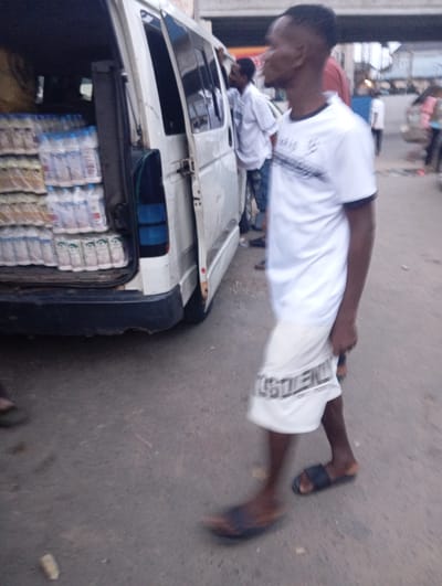 The image depicts a street scene in Port Harcourt, Nigeria. In the foreground, a man wearing a white t-shirt with a black collar and a white patterned skirt is walking. His t-shirt has a black design across the chest. He is wearing dark-colored sandals. Behind him, a white van is parked, with its rear doors open, revealing stacks of bottled water. The van appears to be a commercial vehicle used for distribution. Other individuals are visible in the background, suggesting a public area with some pedestrian and vehicle traffic. The environment appears to be an urban street, with paved road surfaces and buildings in the distance. The lighting suggests it is daytime, possibly late afternoon.