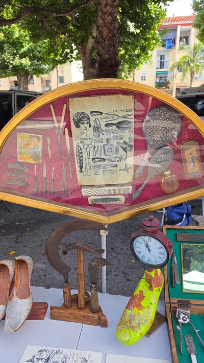 The image captures an outdoor market scene in Almuñécar, Spain. In the foreground, a collection of vintage items is displayed on a table. Dominating the center is a large, ornate gold-trimmed fan-shaped glass case, filled with antique hair styling tools and accessories. These include various combs, scissors, hairpins, a curling iron, a hairbrush, and a decorative hand mirror. A vintage advertisement, featuring a woman with a 1920s hairstyle, is also prominently displayed within the case.
Below the display case, more items are arranged on a white tablecloth. These include a pair of ornate silver women's shoes, a display of vintage metal tools that appear to be agricultural or manual labor implements, a red antique clock, and a brightly painted object resembling a shoe last or wooden mold, decorated with autumn leaves. To the right, a green velvet-lined box holds additional grooming tools, including a straight razor and a hair clipper.
The background of the image shows a street scene typical of a Spanish town, with buildings, trees, and a glimpse of parked vehicles. The lighting suggests it is daytime, with dappled sunlight filtering through the trees. The overall impression is a market stall featuring a curated collection of vintage and antique items, likely for sale.