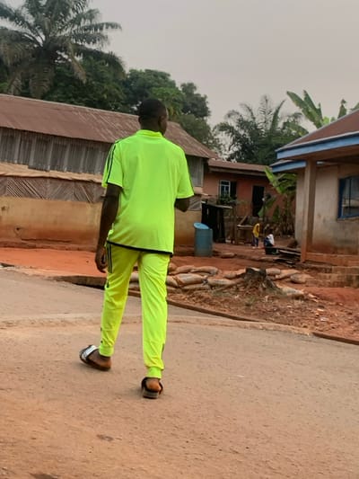 A man in a bright yellow tracksuit stands on a road in front of some buildings. The buildings appear simple, with one showing signs of wear. A pile of firewood or logs rests beside one of the buildings. The sky is overcast, and the environment suggests a rural or less developed area. Palm trees are visible in the background. The man is wearing sandals.