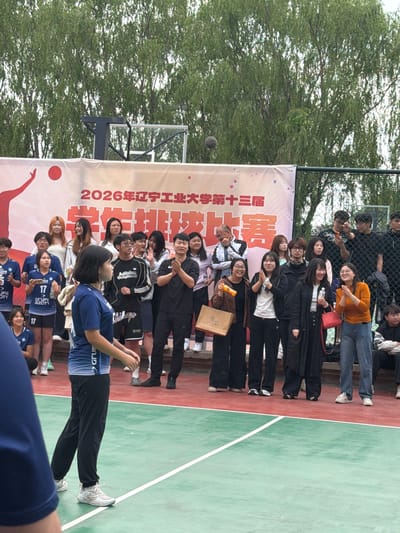Here is a description of the image:
The image appears to be taken outdoors, possibly at a sports facility in Guta, China. The location has a basketball court and what looks like a banner. The banner has Chinese characters and what appears to be the year "2026". It also has a graphic that looks like a volleyball player in action. A group of people is gathered around the banner, most of them looking at the camera. Some are smiling. Several of them are wearing uniforms. A volleyball seems to be in the air, apparently in the process of being served.