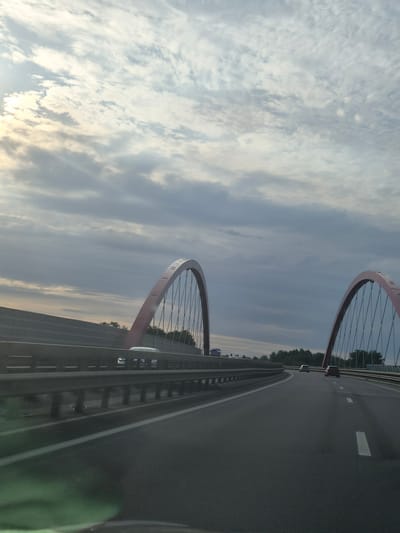 The image shows a view from a vehicle traveling on a highway in Schifflange, Luxembourg, passing under two large, modern red arched bridges. The sky above is cloudy with patches of sunlight breaking through, suggesting either early morning or late afternoon. The highway has multiple lanes marked with white lines. On the left side of the road, a sound barrier runs parallel to the highway. Several cars are visible on the road, some in the distance and some closer. The right side of the highway is lined with trees. The overall scene depicts a contemporary infrastructure setting in an urban or suburban environment.
