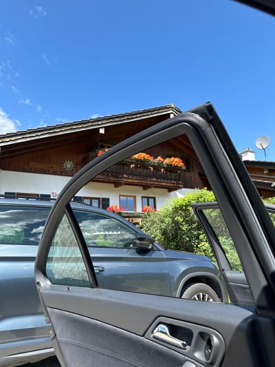 This is a medium shot of a grey car parked in front of a wooden house with a white facade and a satellite dish on the roof, under a clear blue sky with sparse clouds. The house has a balcony with flower boxes filled with orange flowers. A sign on the house reads "HOCHWOHNUNGEN". The left rear door of the car is open, and the interior of the car door is visible in the foreground. The scene is likely in Bischofswiesen, Germany. There are no people visible in the image.