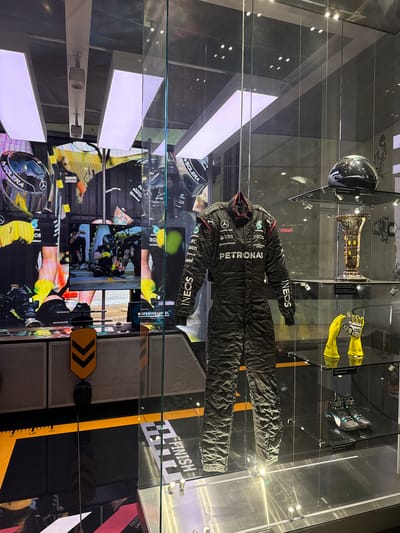 The image shows a display case in Stuttgart, Germany, exhibiting racing memorabilia. In the center, a black Mercedes-AMG Petronas Formula One racing suit is displayed. To the right, a trophy and a pair of bright yellow racing gloves are visible on a shelf. Below these, a pair of black and turquoise racing shoes are displayed. A black racing helmet is positioned on the top shelf. The display case is behind a glass partition, and reflections of lights and the surrounding environment are visible on the glass. In the background, large screens display images of Formula One pit stops and drivers. The overall environment appears to be a museum or exhibition space dedicated to motorsports.