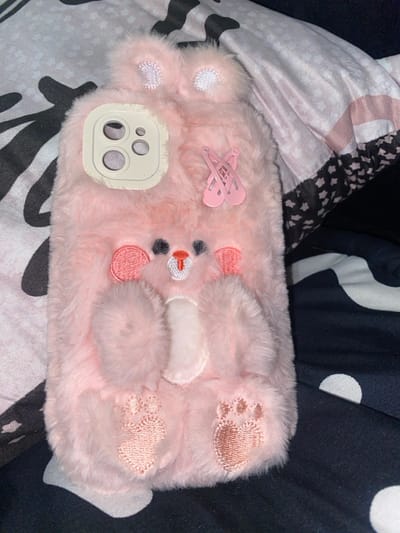 A pink, furry phone case resembling a bear lies on a patterned surface. The case features embroidered details like paw prints, a white belly, and a bear face with pink cheeks and a small orange nose. A white camera cutout is visible on the left side, and two pink hair clips are positioned on the upper left. The background includes a mix of white and black patterned fabric.