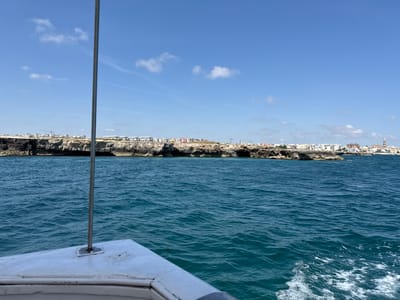 The image presents a view from the bow of a boat looking towards the coastline of Monopoli, Italy, under a bright blue sky. In the foreground, the white, slightly weathered bow section of a vessel is visible, with a vertical metallic pole affixed near its center, extending upwards through the frame. The midground is dominated by choppy, deep blue-green seawater, exhibiting ripples and small waves across its surface, with a faint white wake visible in the lower right corner.

The background features a rugged, rocky coastline forming the horizon. From left to right along the coast, various structures are present. On the far left, light-colored buildings, some appearing new or under construction, sit atop the cliffs, with at least one tall construction crane visible against the sky. Further right, a stretch of rocky shore leads to a more densely built area composed of numerous white and light-colored buildings. Towards the far right, a prominent light-colored tower with a pointed or domed top, indicative of a church spire or bell tower, rises distinctly above the surrounding urban landscape, characteristic of Monopoli's historic center. Sparse green vegetation is visible on parts of the cliffs. A few small, white cumulus clouds are scattered across the otherwise clear blue sky, which occupies the upper half of the frame. One or two indistinct human figures are discernible on a rocky outcrop near the water's edge in the mid-right background.