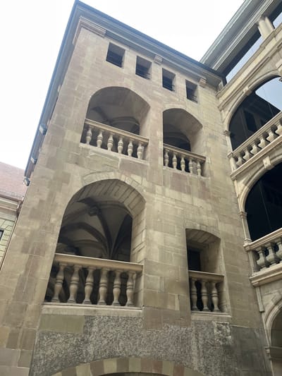 This rotated image captures a detailed section of a historic stone building in Genève, Switzerland, viewed from a low angle looking upwards towards the sky. The main subject is a large, light-colored stone facade characterized by intricate architectural features.
The building exhibits several arched openings. Two prominent, large arched windows in the mid-ground reveal dark interiors with visible ribbed vaulting, suggesting a complex interior structure like a hall or chapel. Each of these large arches is partially framed by a stone balustrade featuring multiple turned balusters. Above and to the left of these, a series of smaller, rectangular windows are set into the stone. The stonework is precise, with visible blocks and mortar lines.
Above the main facade, more elaborate stone architectural elements, including arches and balustrades, are visible against a bright, clear sky, which occupies the upper and left portions of the frame. A dark structural element, possibly an upper level or roof, is also present against the sky. A small portion of a red brick building is visible in the bottom right corner.
The scene is static, devoid of people or any activity. The clear, bright sky indicates it is daytime. No text is visible within the image.