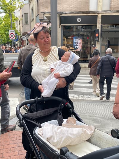 The image captures a street scene in Terrassa, Spain, likely taken during a public event or gathering. The primary subjects are a woman holding a baby, who appears to be sleeping, next to a baby stroller. The woman is wearing a hat with a bow and a dress with a large brooch, and a string of pearls. The baby is dressed in a white knitted outfit with a bonnet. They are situated on a sidewalk with a crosswalk and other pedestrians visible in the background.
Behind them, buildings with shops can be seen, including one with a display advertisement. There is a street sign that shows the speed limit "30". The surrounding environment includes street lights, trees, and a crowd of people in the distance. The overall impression is a moment captured during a daytime event.