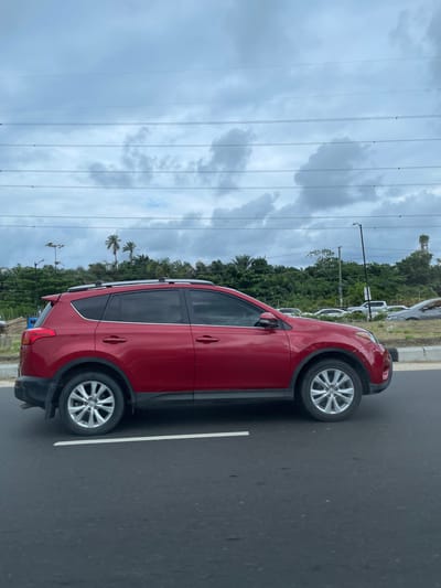 A red SUV is captured in a side profile view, traveling on a paved road. The vehicle features roof rails and tinted windows. The road is marked with a white line. In the background, lush green trees and vegetation line the horizon under a cloudy, overcast sky. Several power lines are visible crisscrossing the sky. Other vehicles are present in the distance on what appears to be a highway or major road. The overall scene suggests an outdoor setting, likely a suburban or roadside environment.