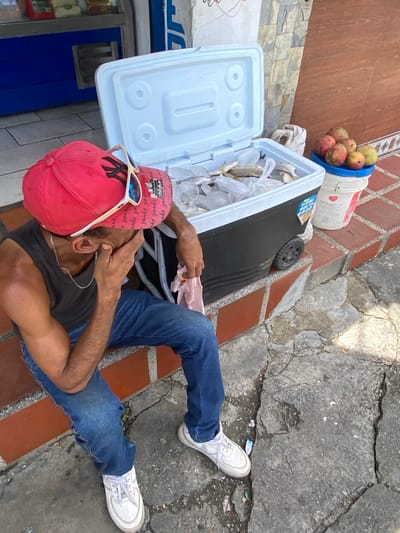 A man sits on a raised brick ledge, his face partially obscured by his hand, wearing a red baseball cap, a dark sleeveless shirt, and blue jeans. In front of him, an open cooler filled with ice and fish rests on the ledge. To the right of the cooler, a blue bucket topped with a white bucket holds several mangoes. The scene appears to be on a street in San José de Barlovento, Venezuela, with a building facade and tiled floor visible in the background.