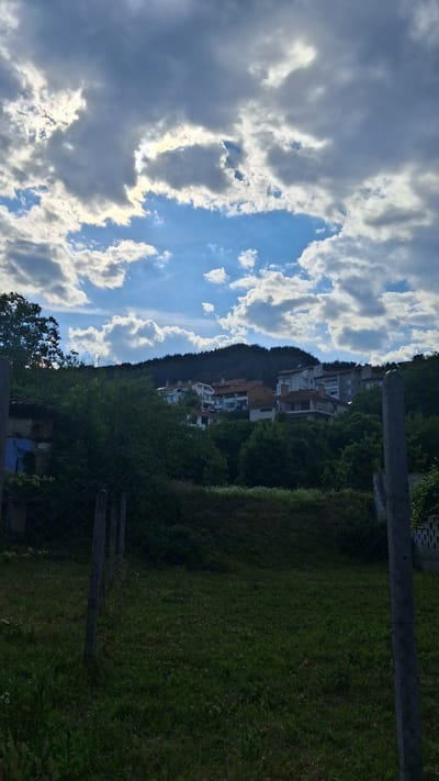 The image shows a view of buildings on a hillside under a cloudy sky. The sky is a mix of blue patches and dramatic grey and white clouds, with sunlight breaking through in some areas. Below the sky, a green, tree-covered hillside is visible, with several buildings clustered on it. These buildings appear to be residential, with a mix of architectural styles, including tiled roofs and multi-story structures. In the foreground, there is a grassy area with a wire fence and concrete posts. The overall impression is of a somewhat overcast but potentially sunny day in a residential area on the outskirts of a town. Given the context, this could be a scene in Gotse Delchev, Bulgaria, depicting a typical residential neighborhood situated on the slopes surrounding the city. No people are visible in the image.