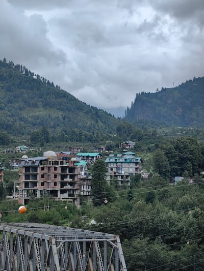 A cloudy, overcast day in a hilly, forested area with buildings and a bridge. The image captures a panoramic view of a town nestled among mountains. In the foreground, a grey metal truss bridge spans across the scene. Behind the bridge, lush green trees cover the lower slopes of the mountains. Several buildings are visible, ranging from unfinished structures to completed homes and hotels with distinctive green roofs. One building under construction is prominently featured, showing its brickwork and incomplete floors. An orange and white spherical object hangs from wires above the bridge. The upper part of the image is dominated by a dramatic cloudy sky, with some portions of the mountains showing steep, rocky cliffs. The location is identified as the city of Kullu, India. There are no people visible in the image.