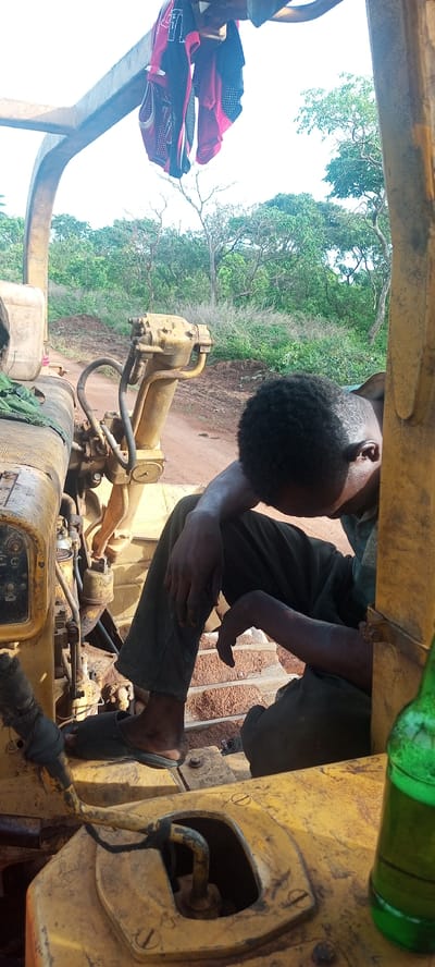 A young man is resting inside the cabin of a large, yellow piece of heavy machinery, likely a bulldozer or similar construction vehicle. He is seated on the right side of the frame, leaning forward with his head down, appearing to be asleep or deeply contemplative. His dark, curly hair is visible, and his skin is dark. He wears dark pants and black flip-flops. His hands and arms show signs of dirt or grime, suggesting manual work.

The machinery itself is old and robust, painted yellow, with various levers, pipes, and gauges visible on the console to the left of the man. Faint labels like "OFF", "WASH", and "RESET" can be partially discerned on some controls. Above the man's head, two pieces of red and black athletic-style clothing, possibly jerseys, are hung to dry or air out from an overhead bar of the vehicle's frame. To the far right in the foreground, a green glass bottle is visible, resting on a part of the machinery.

The setting is outdoors, under bright daylight, indicated by the strong shadows and clear sky. In the background, a dirt road or path stretches into the distance, flanked by lush green trees and bushes, suggesting a rural or construction site environment. The ground visible behind the man and the machinery is reddish-brown, typical of laterite soil. The overall impression is one of a break from labor in a warm, outdoor environment in Otunade, Nigeria.