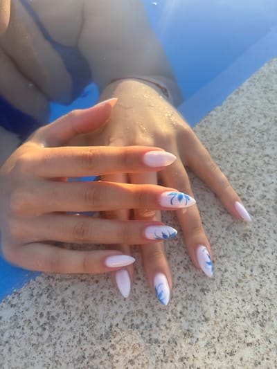 A close-up, overhead shot captures a person's hands and nails submerged in clear blue water, resting on a textured, light-colored surface. The water is illuminated by golden sunlight, creating bright highlights on the skin and nails. The nails are long and almond-shaped, with a light pink or white polish. Some nails feature delicate blue floral or abstract designs. A thin red string bracelet is visible on the wrist. The textured surface appears to be the edge of a pool or a similar structure, made of speckled stone. The overall scene suggests a leisurely moment by the water.