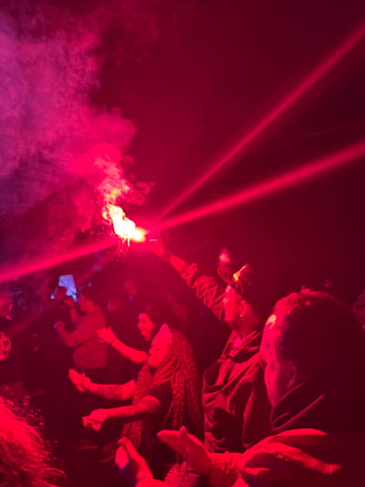An indoor gathering in Большая Кивара, Russia, is depicted under intense red artificial lighting and significant atmospheric smoke. In the central-left, an individual holds a burning orange-yellow flare, which emits a bright flame, sparks, and dense smoke that diffuses into the environment. Two distinct, diagonal red laser beams traverse the upper right portion of the frame. Numerous people are present, largely obscured by the low light and red illumination. In the mid-right, a woman with long, braided hair and visible arm tattoos is captured with her arms raised, suggesting movement or dancing. Other figures throughout the scene display similar gestures, with some individuals in the left-midground seen holding mobile phones, one of which has a glowing blue-white screen, possibly recording the event. The scene conveys a dynamic social gathering or performance.