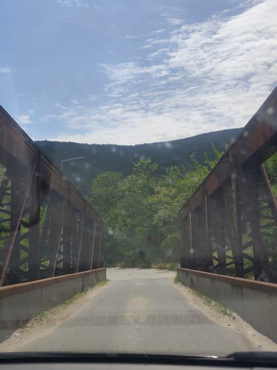 The image shows a narrow asphalt road leading onto an old, rusty, metal truss bridge in Jeleznitsa, Bulgaria. The bridge appears to be a single lane. The sides of the bridge consist of low concrete barriers topped with metal railings. Beyond the bridge, trees and vegetation obscure the landscape. A hill is visible in the background. The sky is partly cloudy. A street light is observed off to the left side. The image is taken from the perspective of someone in a vehicle, with the dashboard partially visible at the bottom.