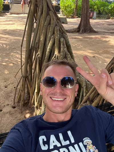A man is taking a selfie in a hammock, possibly at Bang Niang Beach in Ban Khuk Khak, Thailand. He is wearing sunglasses and a navy blue t-shirt with graphics on it. He is smiling and holding up two fingers in a peace sign. The background shows sand, large tree roots, and lush green vegetation. In the distance, there are stairs and a trash can. The man appears relaxed and is enjoying the tropical environment.